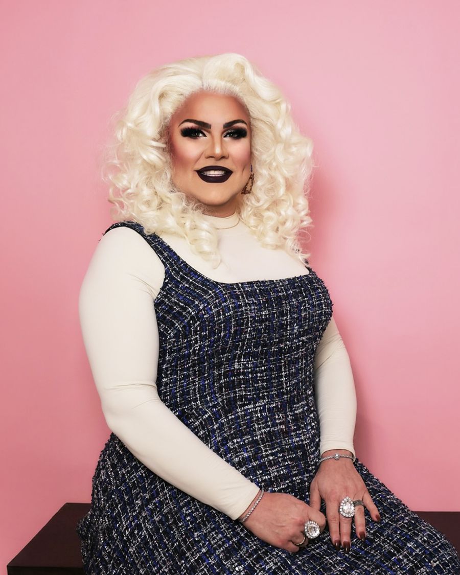Abby Cummings seated in a tweed dress and cream turtleneck, platinum curls, smiling against a blush backdrop